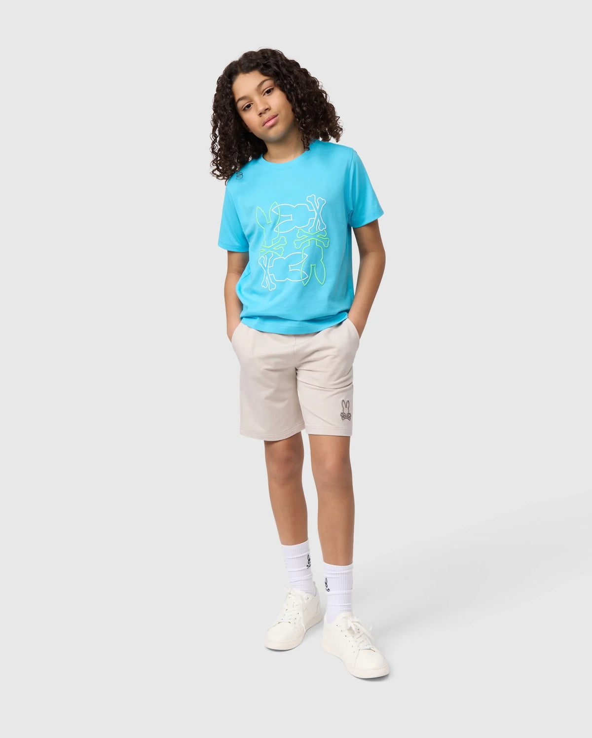 A young girl stands confidently with one hand in her pocket, wearing a blue t-shirt, Psycho Bunny KIDS LIVINGSTON TERRY SWEATSHORT - B0R408B200, white socks, and sneakers against a plain background.