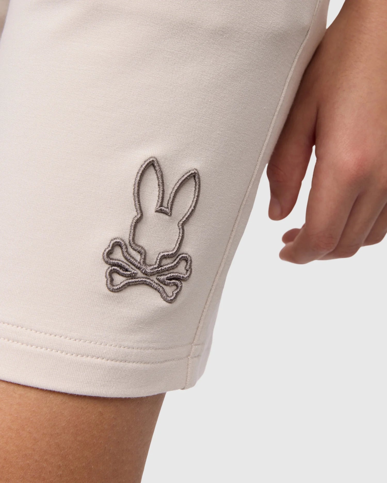 Close-up of a white micro French terry fabric with an embroidered design of a rabbit on the left thigh of a Psycho Bunny KIDS LIVINGSTON TERRY SWEATSHORT - B0R408B200, with a human hand partially visible.