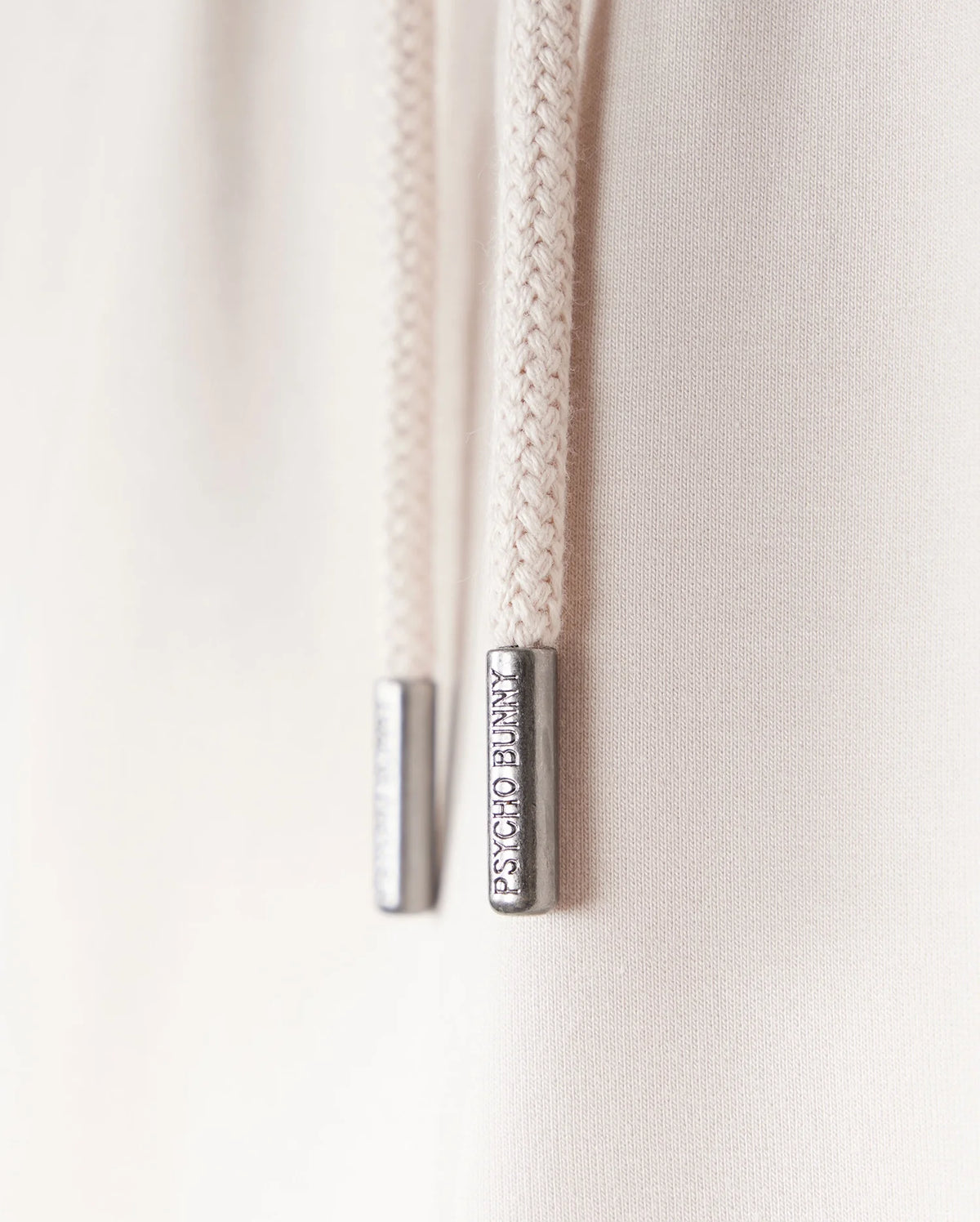 Close-up of two cream-colored drawstring cords with metal tips engraved with "LIMITED EDITION." The cords, part of a Psycho Bunny KIDS LIVINGSTON TERRY SWEATSHORT - B0R408B200, hang against a soft, light background.