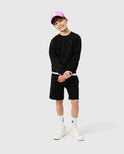 A young boy poses confidently, wearing a black long-sleeve top, Psycho Bunny KIDS LYONS SWEATSHORT - B0R578C200, and white sneakers. He smiles and sports a pink baseball cap, standing against a plain light background.