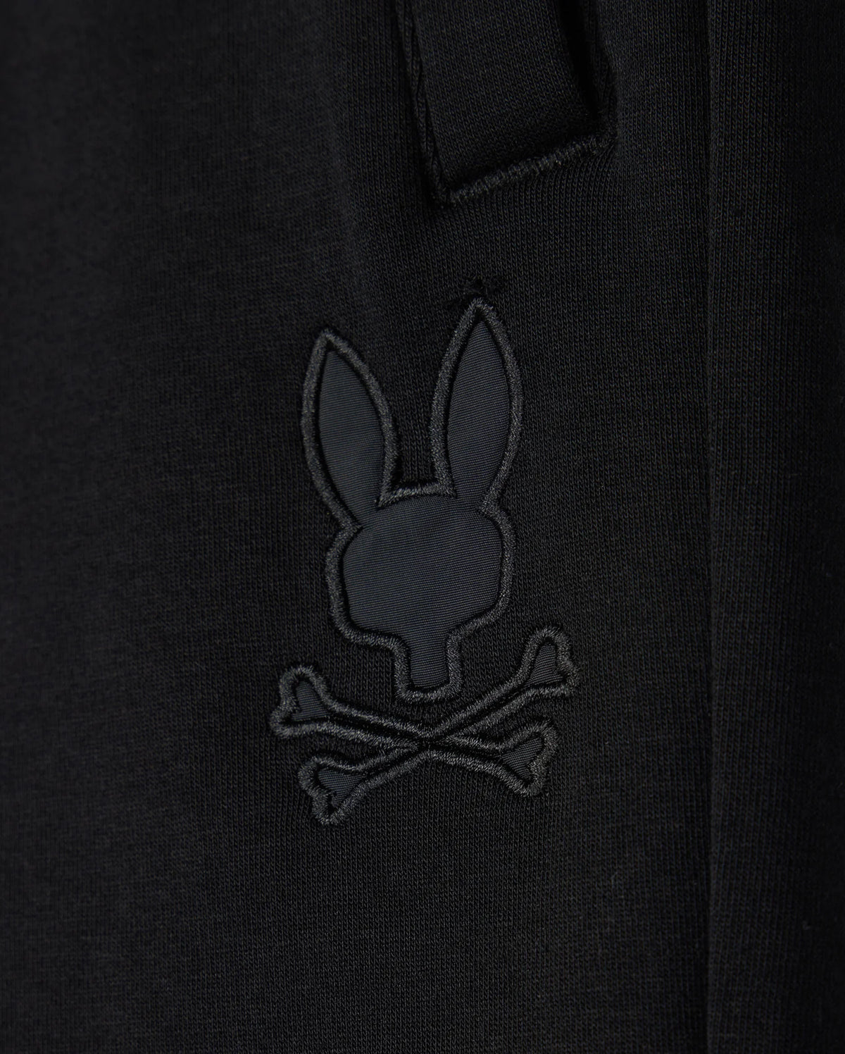 Close-up of a black cotton blend Kids Lyons Sweatshort by Psycho Bunny with an embroidered patch featuring a stylized skull and crossbones design shaped like a rabbit head.