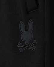 Close-up of a black cotton blend Kids Lyons Sweatshort by Psycho Bunny with an embroidered patch featuring a stylized skull and crossbones design shaped like a rabbit head.