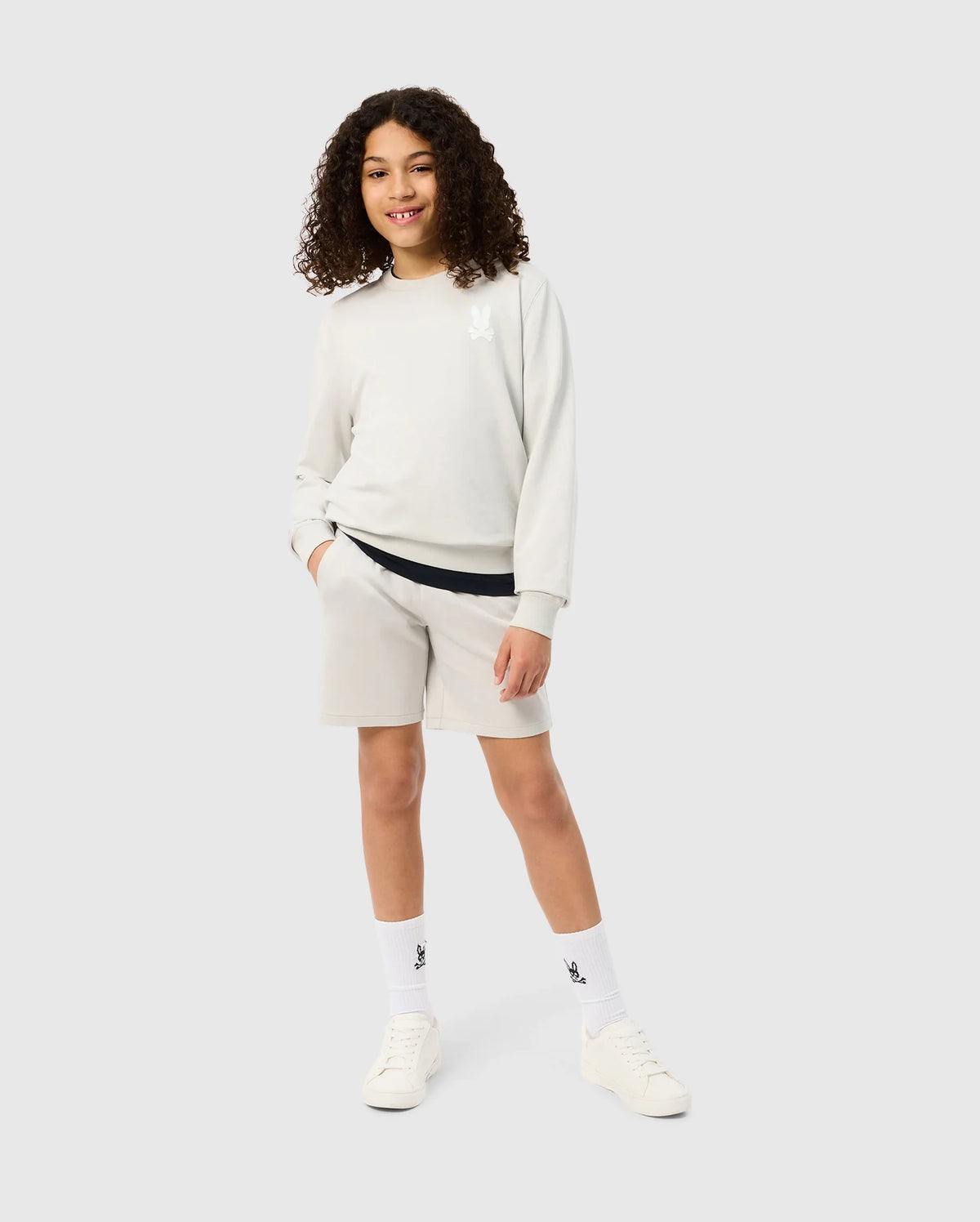 A young person with curly hair smiles while posing in a light-colored sweatshirt and matching Psycho Bunny KIDS LYONS SWEATSHORT - B0R578C200 made from soft stretch modal-cotton-blend. They are wearing white socks and white sneakers. One hand is in the shorts pocket, and they stand against a plain grey background.