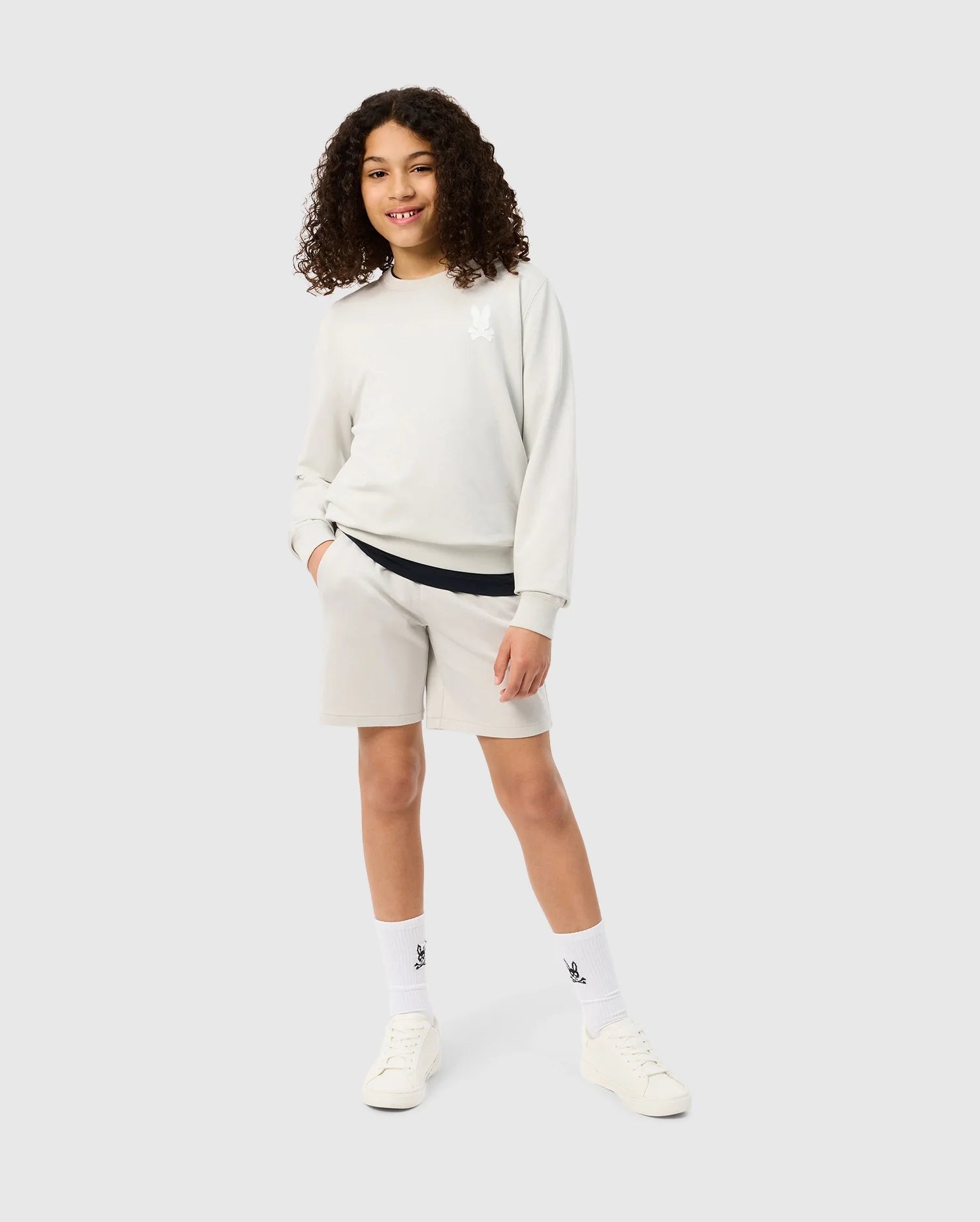 A young person with curly hair smiles while posing in a light-colored sweatshirt and matching Psycho Bunny KIDS LYONS SWEATSHORT - B0R578C200 made from soft stretch modal-cotton-blend. They are wearing white socks and white sneakers. One hand is in the shorts pocket, and they stand against a plain grey background.