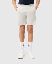 Person wearing beige Psycho Bunny KIDS LYONS SWEATSHORT - B0R578C200, a black t-shirt, white socks with a small black logo, and white sneakers. The person is standing with hands in pockets against a plain white background.