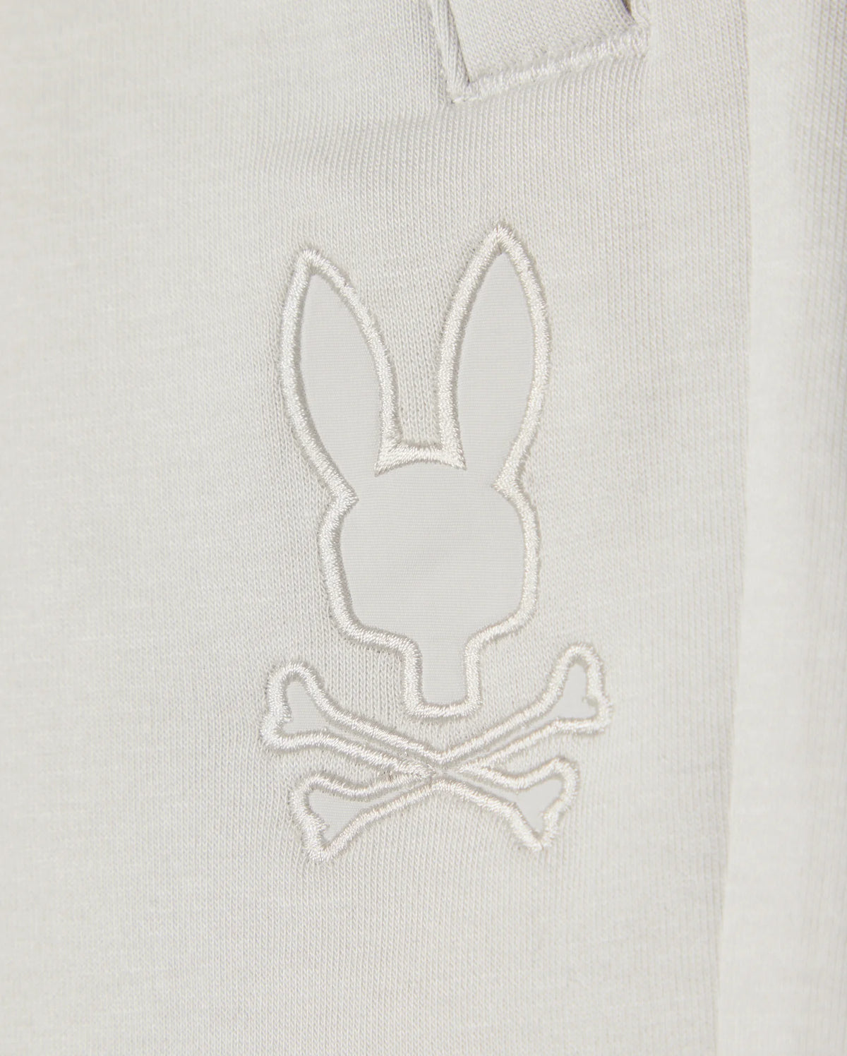 Close-up of a white Kids casual short made from soft stretch modal-cotton-blend jersey, featuring a minimalist embroidered design resembling a rabbit head with crossed bones underneath. The white stitching on the KIDS LYONS SWEATSHORT - B0R578C200 by Psycho Bunny creates a subtle, monochromatic effect.