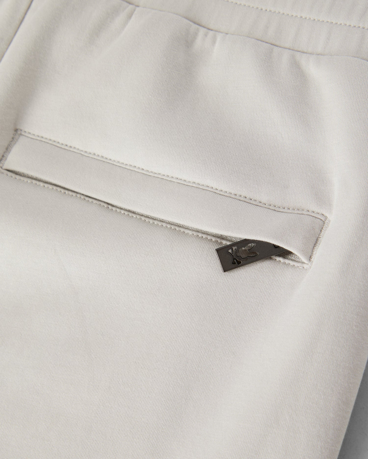 Close-up of the back pocket of a pair of light-colored KIDS LYONS SWEATSHORT - B0R578C200, featuring a small black tag with a subtle design peeking out from the pocket. Made from soft stretch modal-cotton-blend jersey, the fabric appears smooth and slightly elastic. The sweatshorts have a clean and modern look. Psycho Bunny promotes both style and comfort in this garment designed for active kids.
