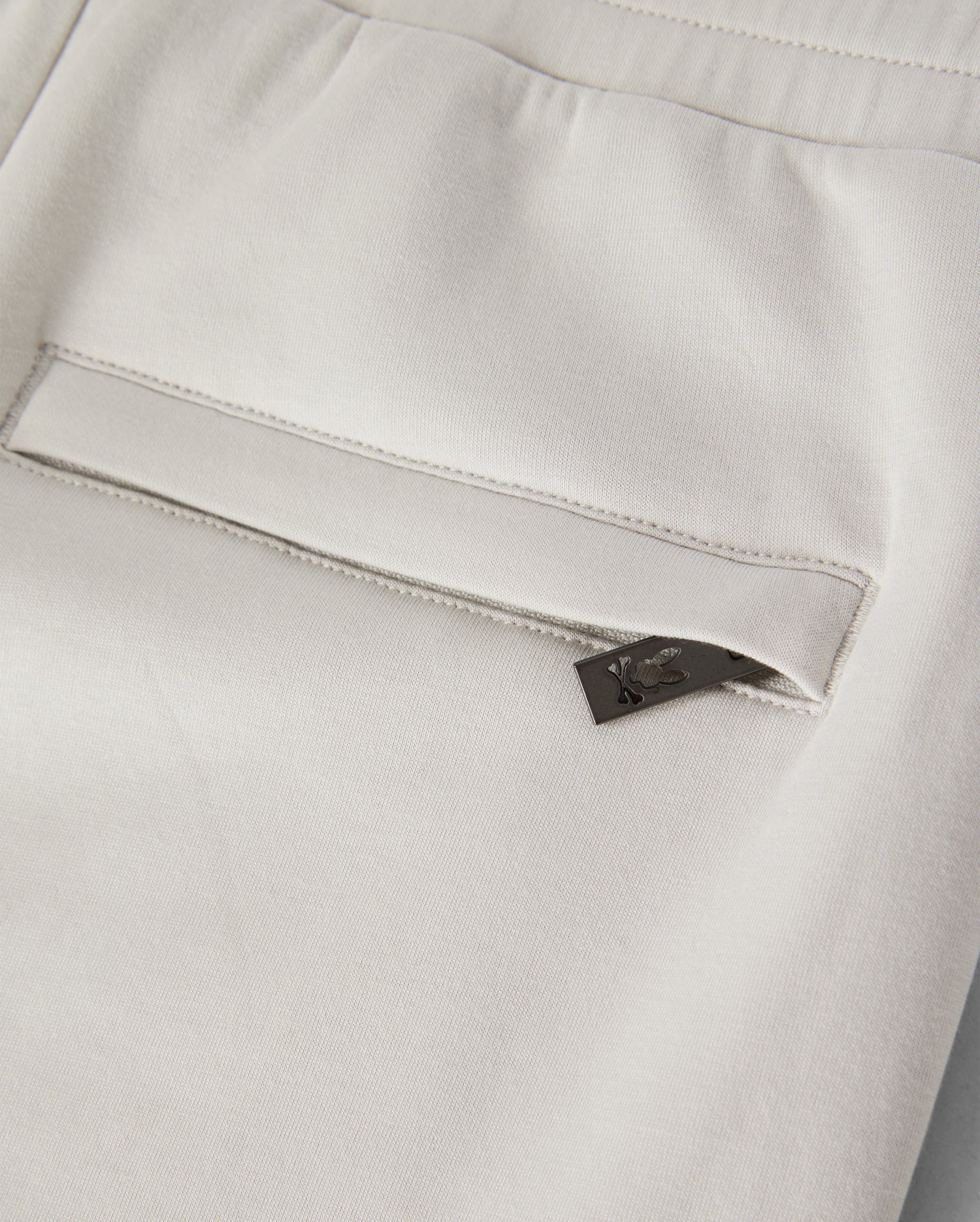 Close-up of the back pocket of a pair of light-colored KIDS LYONS SWEATSHORT - B0R578C200, featuring a small black tag with a subtle design peeking out from the pocket. Made from soft stretch modal-cotton-blend jersey, the fabric appears smooth and slightly elastic. The sweatshorts have a clean and modern look. Psycho Bunny promotes both style and comfort in this garment designed for active kids.