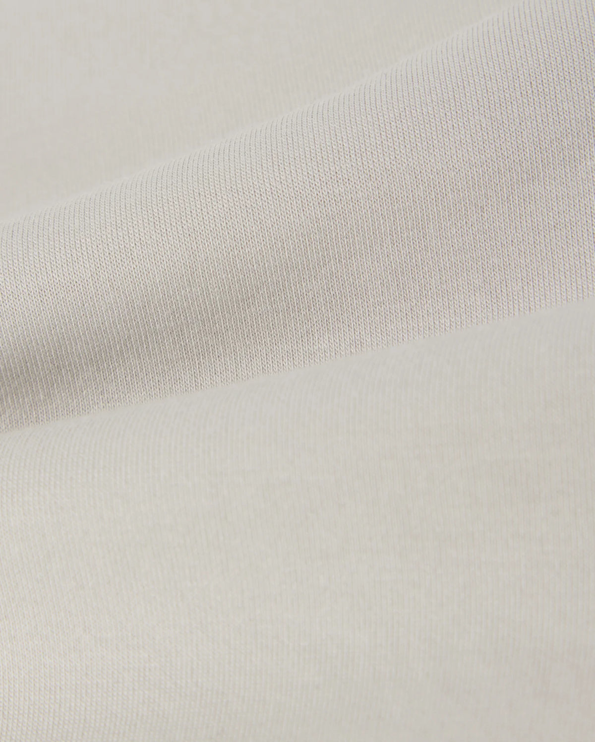 A close-up of a smooth, light beige fabric with a subtle texture. The material shows gentle folds, creating soft shadows and highlights across the surface. This lightweight, finely woven fabric is reminiscent of the soft stretch modal-cotton-blend jersey used in Psycho Bunny's KIDS LYONS SWEATSHORT - B0R578C200 for comfort and style.
