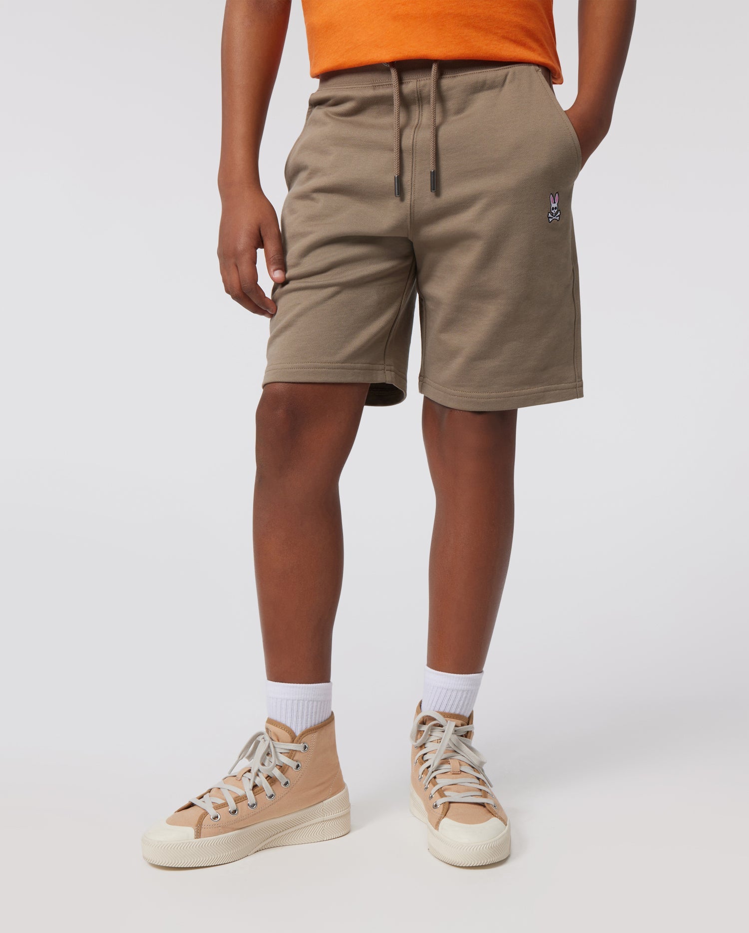 A person is standing against a plain background, wearing an orange shirt, KIDS FRENCH TERRY SHORTS - B0R829ARFT by Psycho Bunny with a small embroidered emblem on the left leg, white socks, and tan high-top sneakers with white laces.