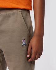 A person wearing Psycho Bunny KIDS FRENCH TERRY SHORTS - B0R829ARFT with a small embroidered logo of a pink bunny and crossbones on the hip. The person is also wearing an orange shirt, and only their lower torso and hand are visible against a plain background.