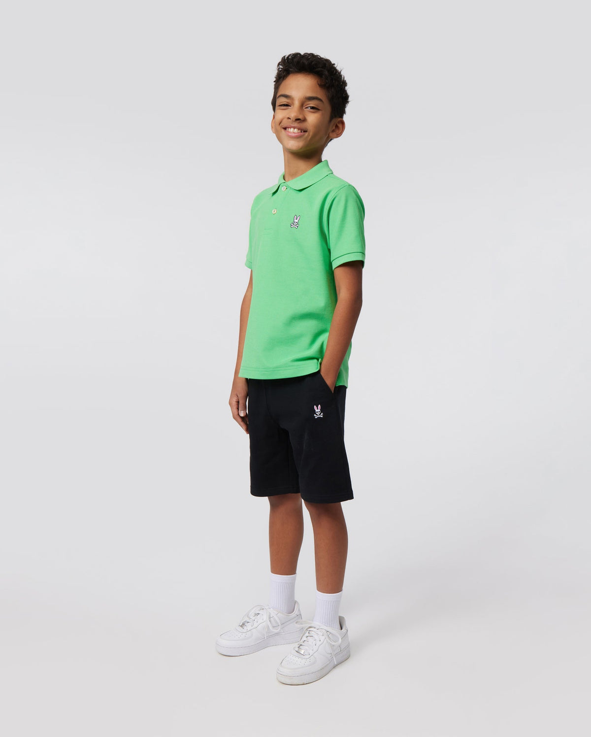 A smiling young boy stands against a plain background, wearing a bright green short-sleeve polo shirt, navy blue KIDS CLASSIC FRENCH TERRY SHORTS - B0R829ARFT by Psycho Bunny with an embroidered Bunny logo, white socks, and white sneakers. He has his hands in his pockets and is looking at the camera.