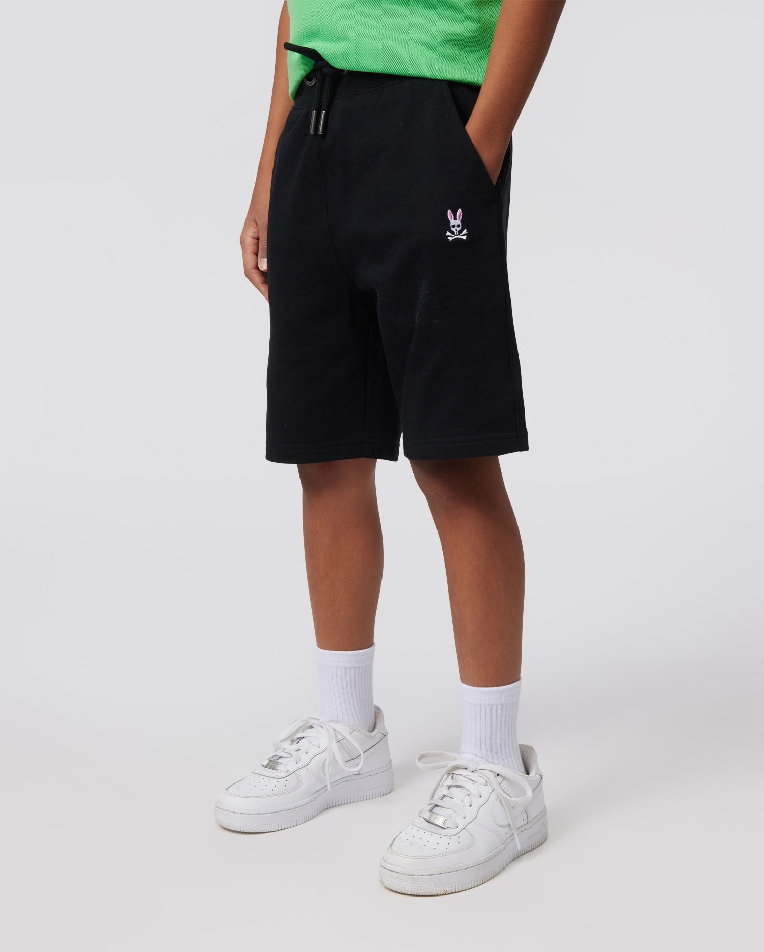 A person is wearing a green shirt, Psycho Bunny KIDS CLASSIC FRENCH TERRY SHORTS - B0R829ARFT with an embroidered Bunny logo on the left thigh, white socks, and white sneakers. The background is plain and light-colored. The person's hands are in their pockets, and their face is not visible.