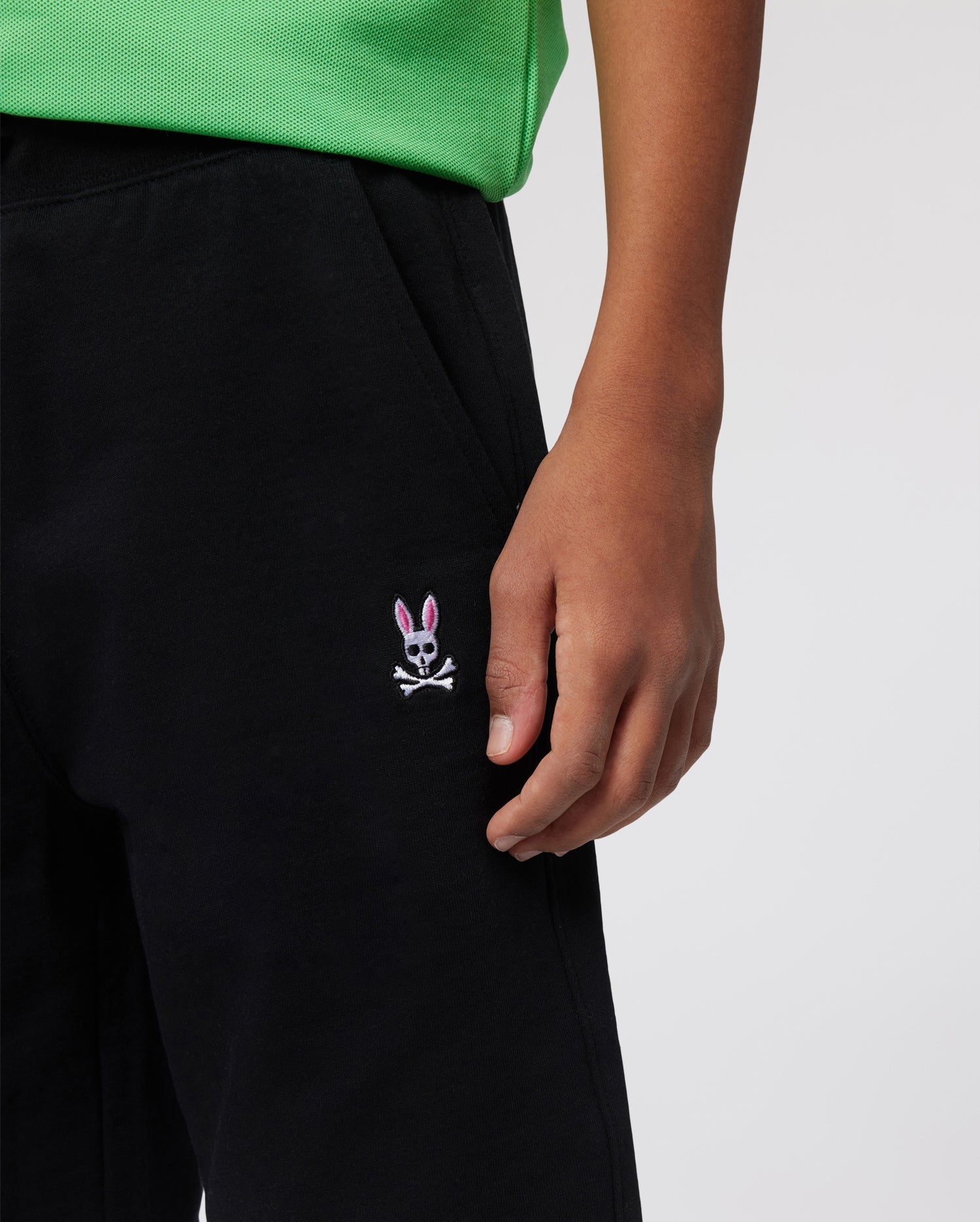 A person wearing a green shirt and black pants. The pants, reminiscent of KIDS CLASSIC FRENCH TERRY SHORTS - B0R829ARFT by Psycho Bunny in their playful design, have an embroidered bunny logo with pink ears and white features positioned near the right pocket. Only the lower torso and hand are visible.