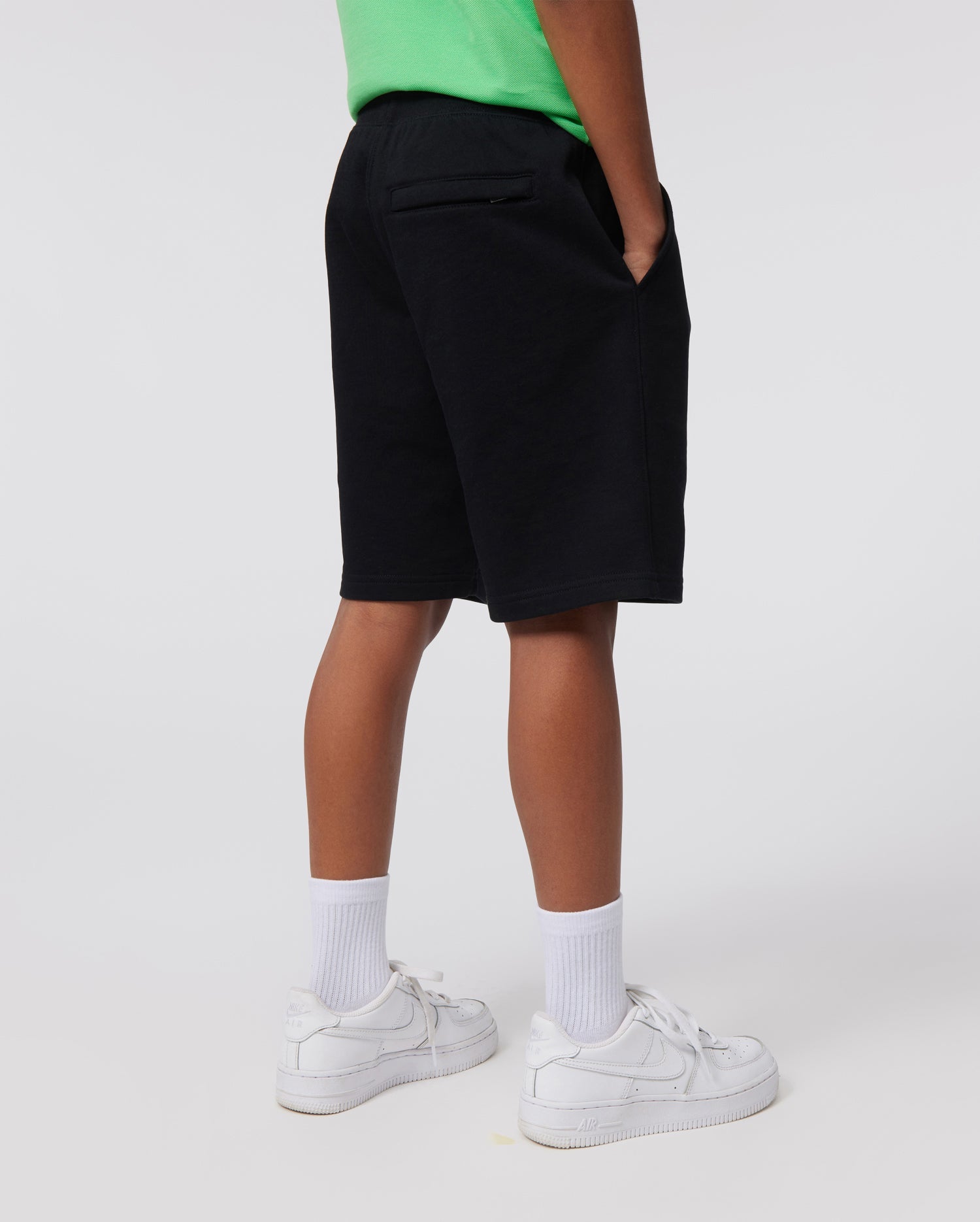 A person wearing a green shirt, white socks, and white sneakers stands with their hands in their pockets. The background is plain and light-colored. The person is shown from the back, focusing on their legs and lower torso. They are also wearing black KIDS CLASSIC FRENCH TERRY SHORTS - B0R829ARFT by Psycho Bunny, featuring an embroidered Bunny logo.
