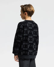 A young boy with short hair stands in profile, dressed in the KIDS ROMEO LIGHTWEIGHT SWEATSHIRT – B0S129E200 by Psycho Bunny. The sweatshirt is black, featuring an enchanting bunny-patterned design. A plain white background accentuates the whimsical details of his cozy attire.