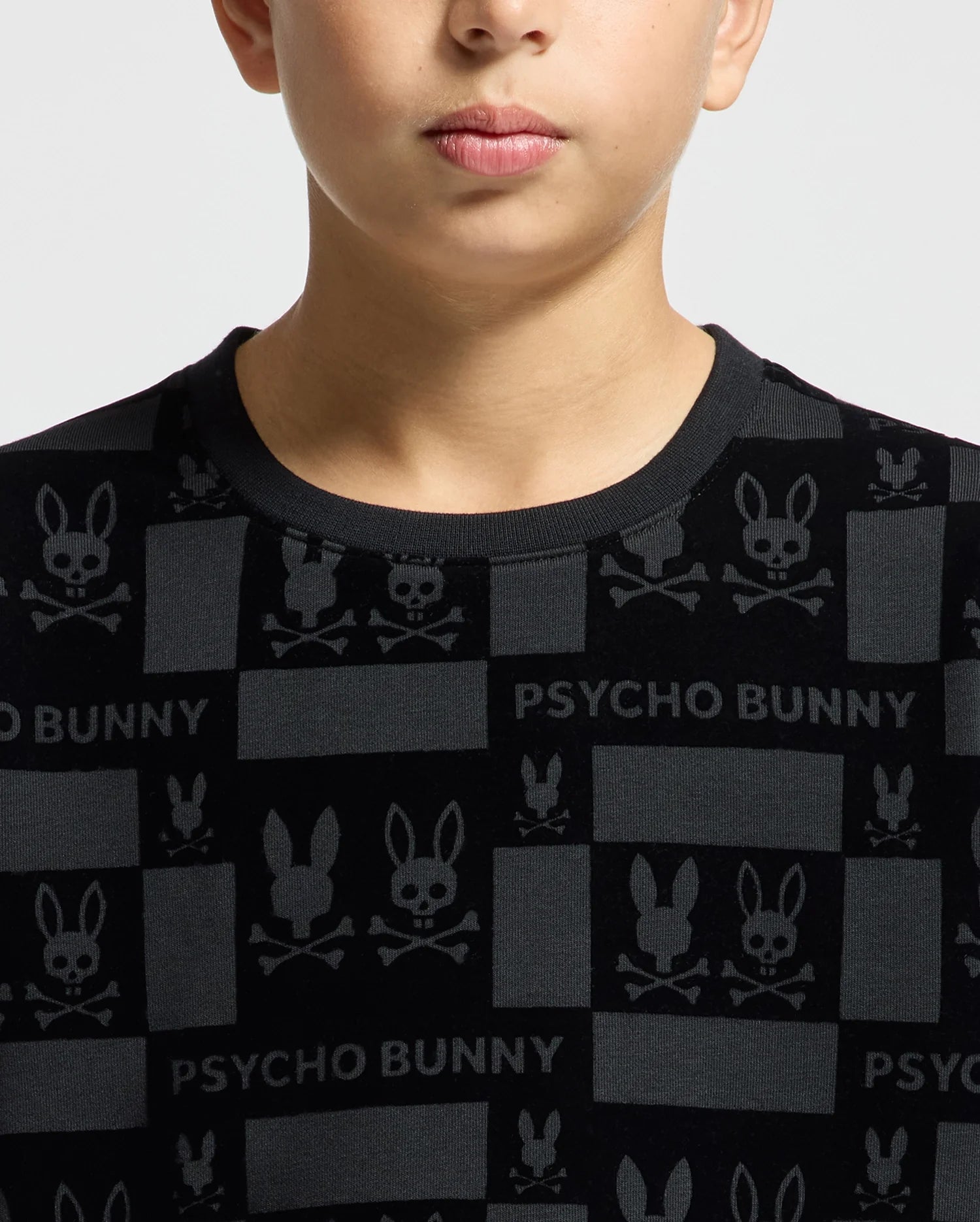 A person is wearing the KIDS ROMEO LIGHTWEIGHT SWEATSHIRT in a black color, featuring a velvet-like texture with designs of skulls, crossbones, and rabbit ears along with the "PSYCHO BUNNY" text. The shirt's grid-themed pattern includes bunny designs in darker tones, adding to its unique style.