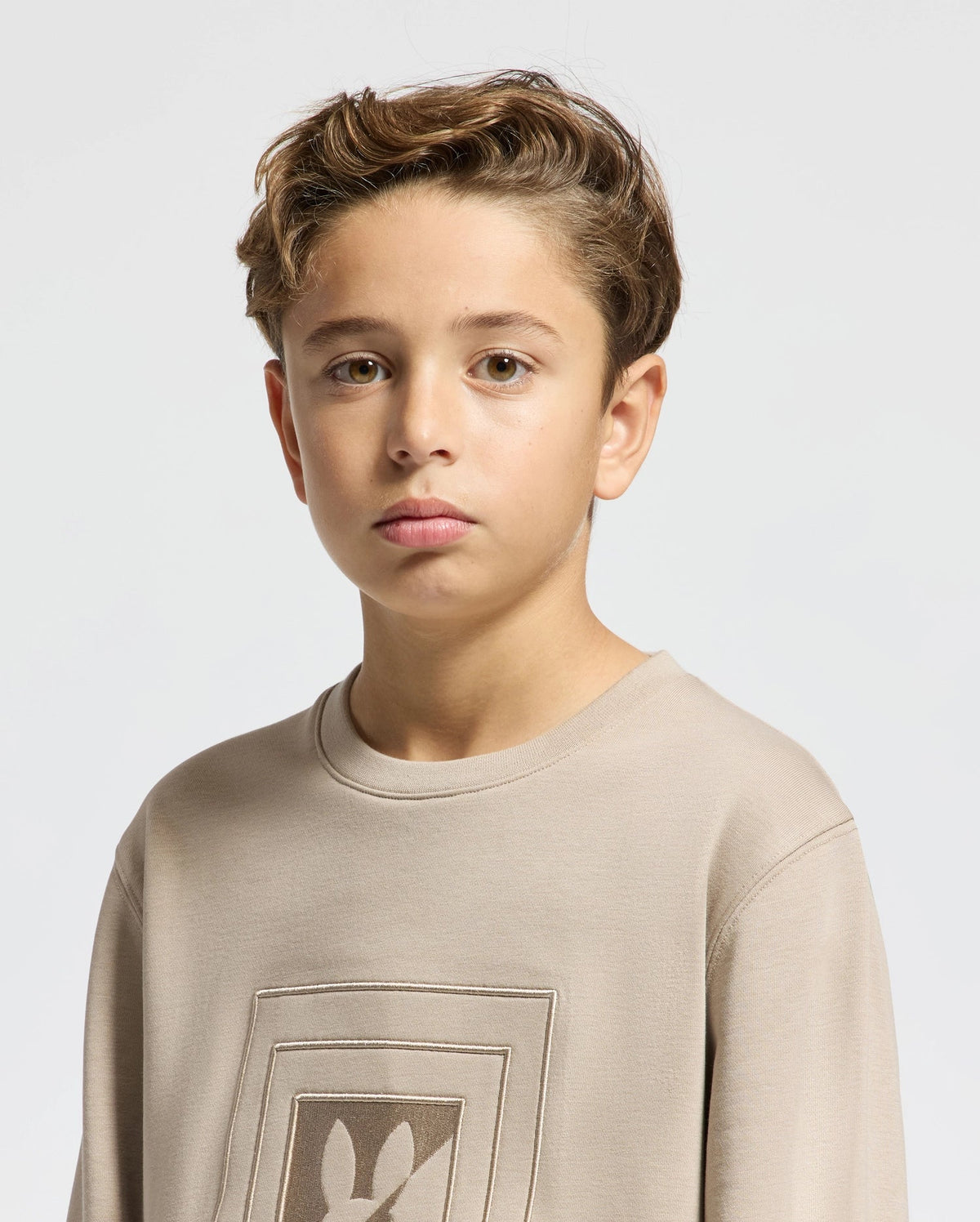 A young boy with short, wavy hair looks directly at the camera. He's wearing the Psycho Bunny KIDS OAK CITY CREW NECK SWEATSHIRT - B0S274E200, featuring a straightforward design perfect for children. The light gray background provides a neutral setting.