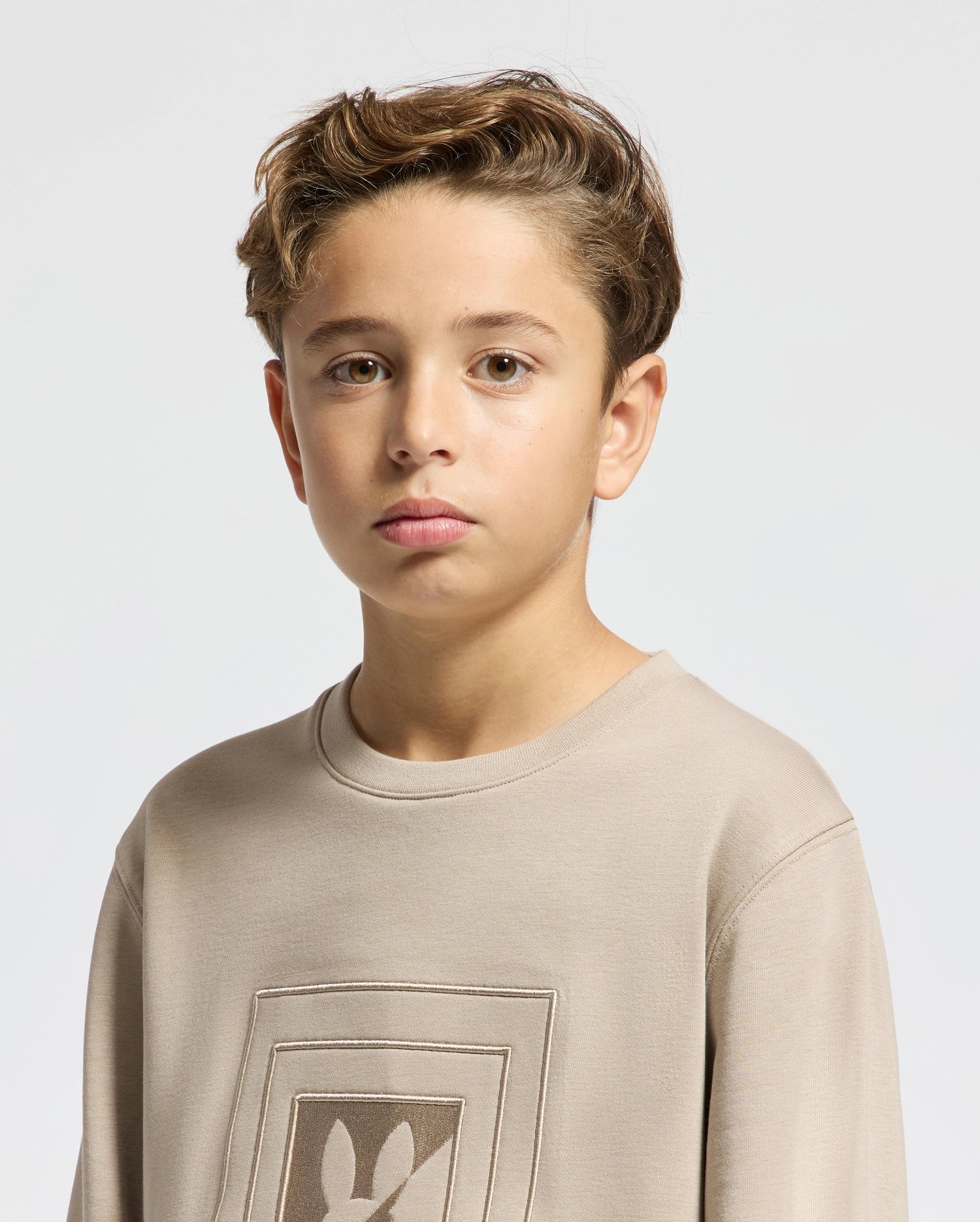 A young boy with short, wavy hair looks directly at the camera. He's wearing the Psycho Bunny KIDS OAK CITY CREW NECK SWEATSHIRT - B0S274E200, featuring a straightforward design perfect for children. The light gray background provides a neutral setting.