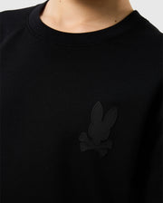 Close-up of a person wearing the Psycho Bunny KIDS HOUSTON FRENCH TERRY CREWNECK - B0S462C200, a black sweatshirt made from premium cotton fabric. It features a subtle black emblem on the chest depicting a stylized rabbit and crossbones, with a lightweight crewneck design. The person's face and background remain out of view.