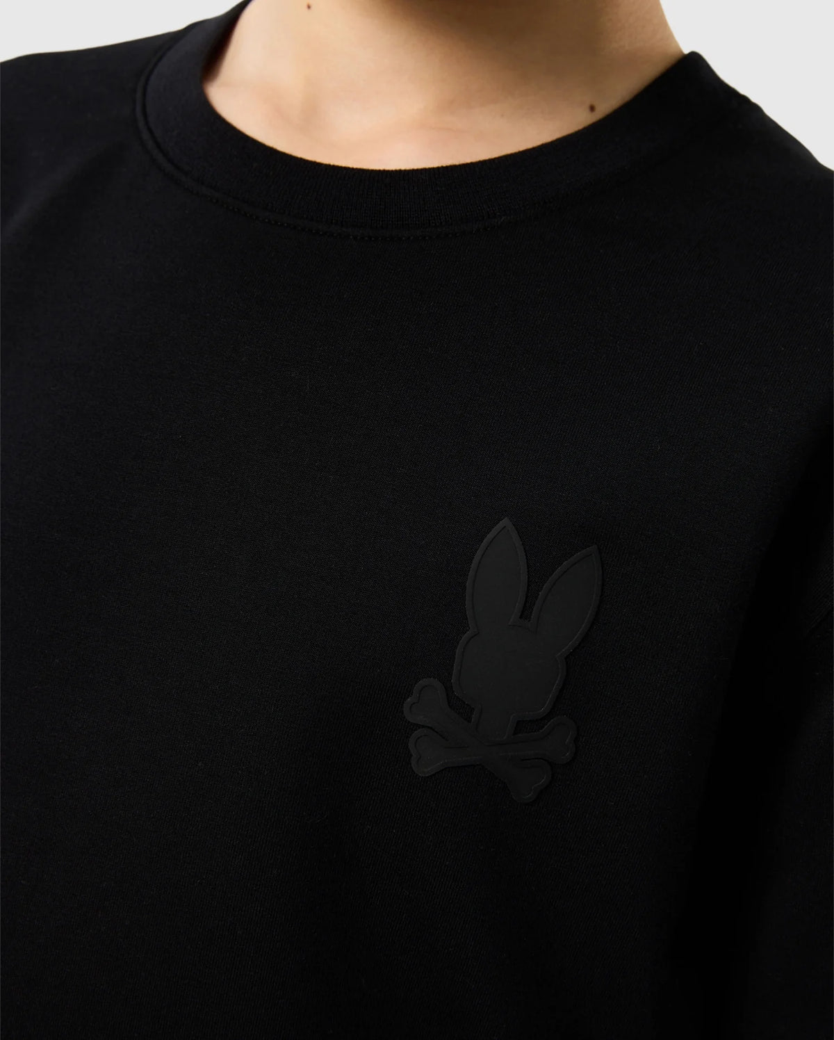 Close-up of a person wearing the Psycho Bunny KIDS HOUSTON FRENCH TERRY CREWNECK - B0S462C200, a black sweatshirt made from premium cotton fabric. It features a subtle black emblem on the chest depicting a stylized rabbit and crossbones, with a lightweight crewneck design. The person's face and background remain out of view.