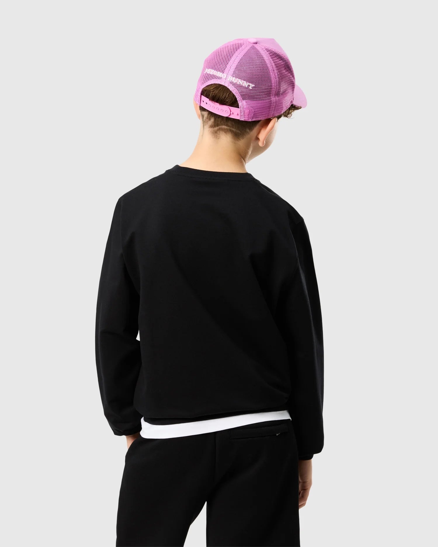 A child wearing a Psycho Bunny KIDS HOUSTON FRENCH TERRY CREWNECK - B0S462C200 and black pants stands with their back to the camera. They are sporting a pink mesh baseball cap, expertly crafted from premium cotton fabric, against a plain gray background.