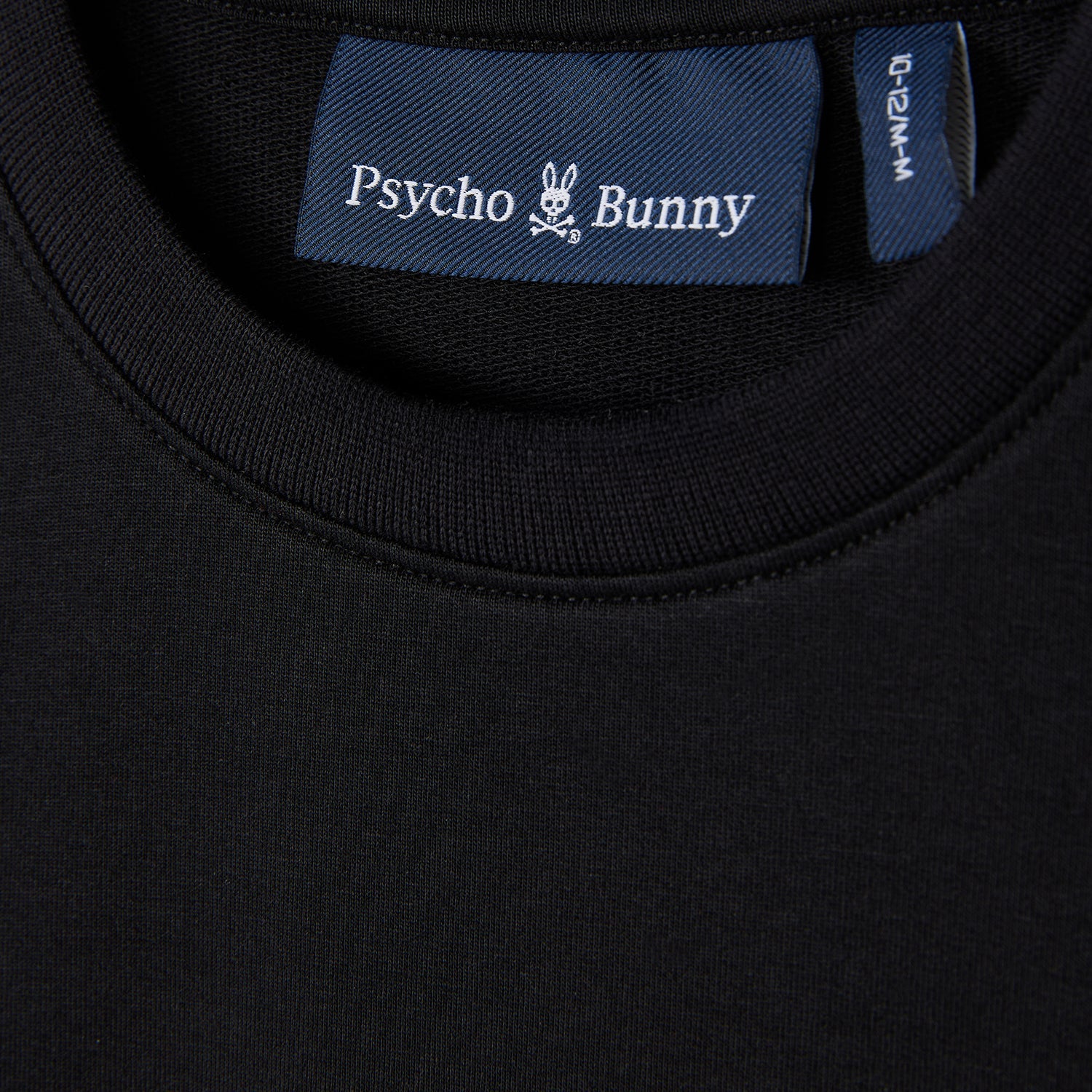 Close-up of a black KIDS HOUSTON FRENCH TERRY CREWNECK (B0S462C200) by Psycho Bunny, showcasing the collar and brand's label inside. Crafted from lightweight Micro French Terry, the label features the signature skull with bunny ears logo, exemplifying the premium cotton fabric quality.