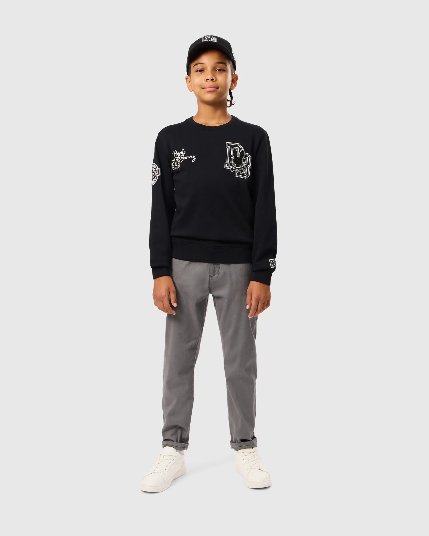A young individual confidently stands against a plain, light gray background, wearing a KIDS ZACK VARSITY SWEATSHIRT by Psycho Bunny in black with chenille embroidery, paired with gray pants, white sneakers, and a black cap.