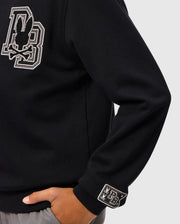 A person is wearing the KIDS ZACK VARSITY SWEATSHIRT (B0S880D200) by Psycho Bunny, featuring a large chenille embroidered letter "B" and a vintage-inspired logo on the chest, along with a smaller logo patch on the wrist. Their hand is tucked into the pocket of gray pants against a plain background.
