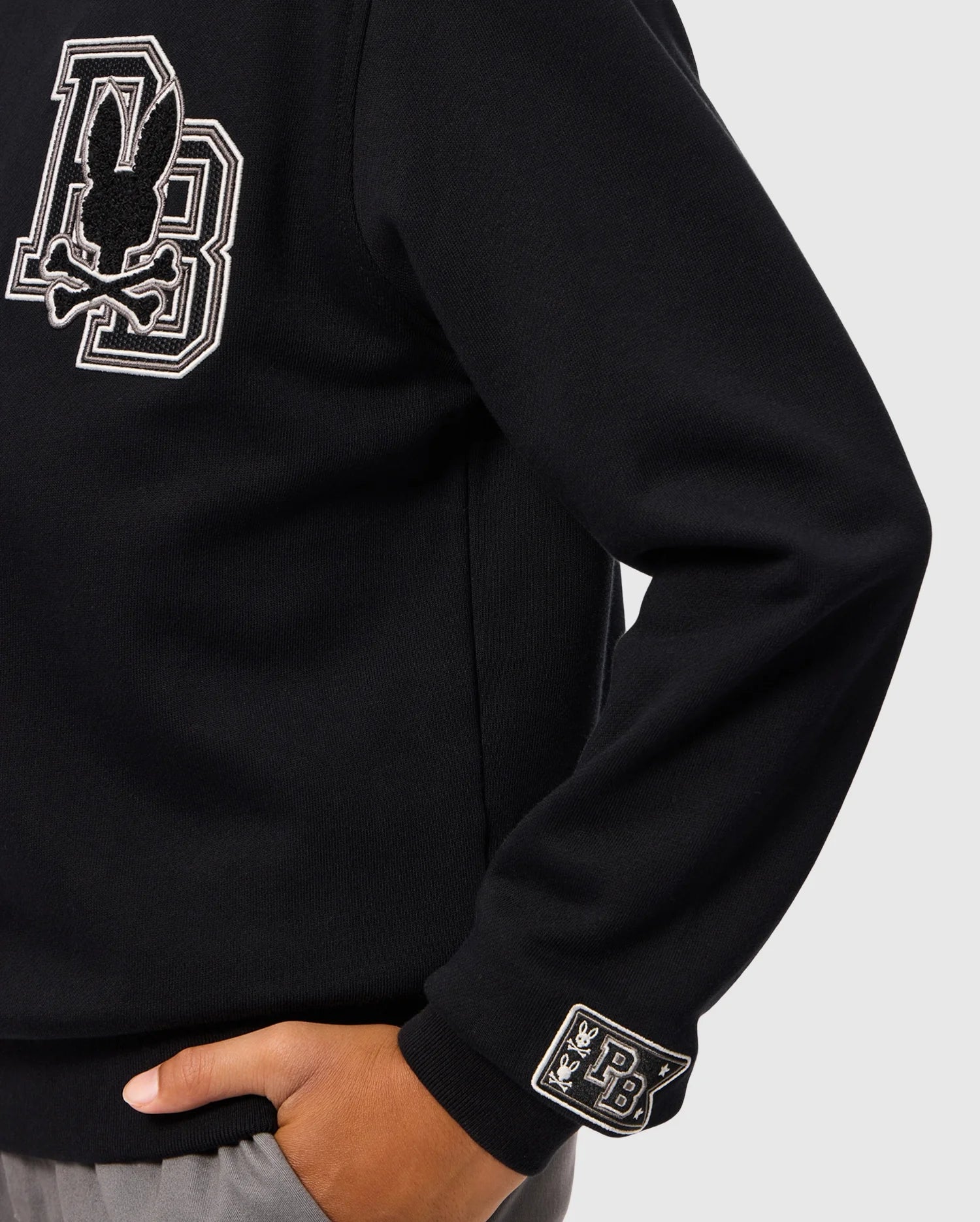 A person is wearing the KIDS ZACK VARSITY SWEATSHIRT (B0S880D200) by Psycho Bunny, featuring a large chenille embroidered letter "B" and a vintage-inspired logo on the chest, along with a smaller logo patch on the wrist. Their hand is tucked into the pocket of gray pants against a plain background.