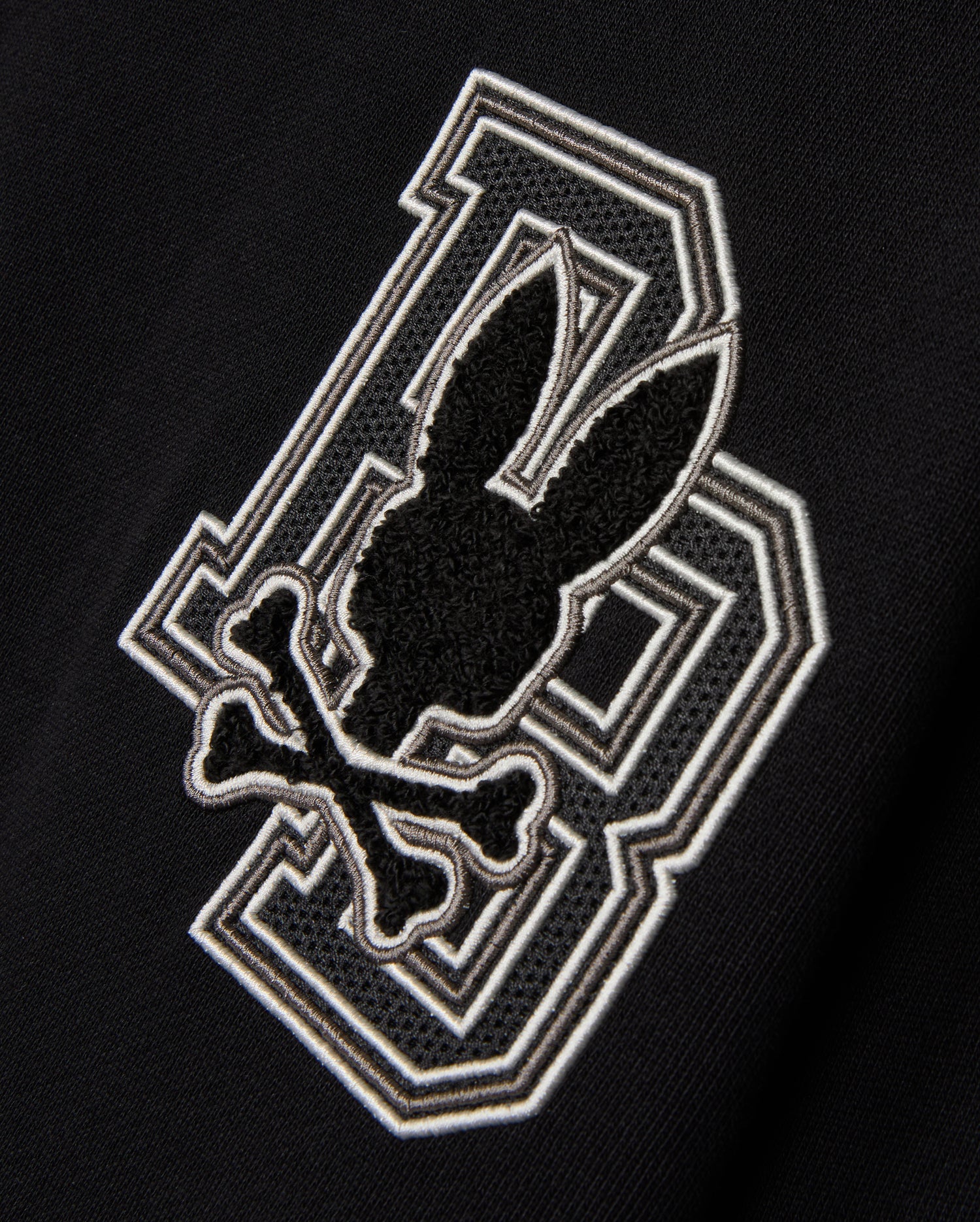 The KIDS ZACK VARSITY SWEATSHIRT by Psycho Bunny features a striking chenille embroidery design with a large letter 'B' outlined in white. Overlapping it is a playful skull and crossbones motif, where a bunny head sits above crossed bones, all in black and white outlines, reminiscent of a vintage-inspired logo.