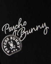 Adorning the black fabric of the KIDS ZACK VARSITY SWEATSHIRT - B0S880D200 by Psycho Bunny, white chenille embroidery spells out "Psycho Bunny." Below it, a vintage-inspired logo displays a circular emblem that surrounds a bunny head with crossbones, evoking the look of a classic varsity sweatshirt.