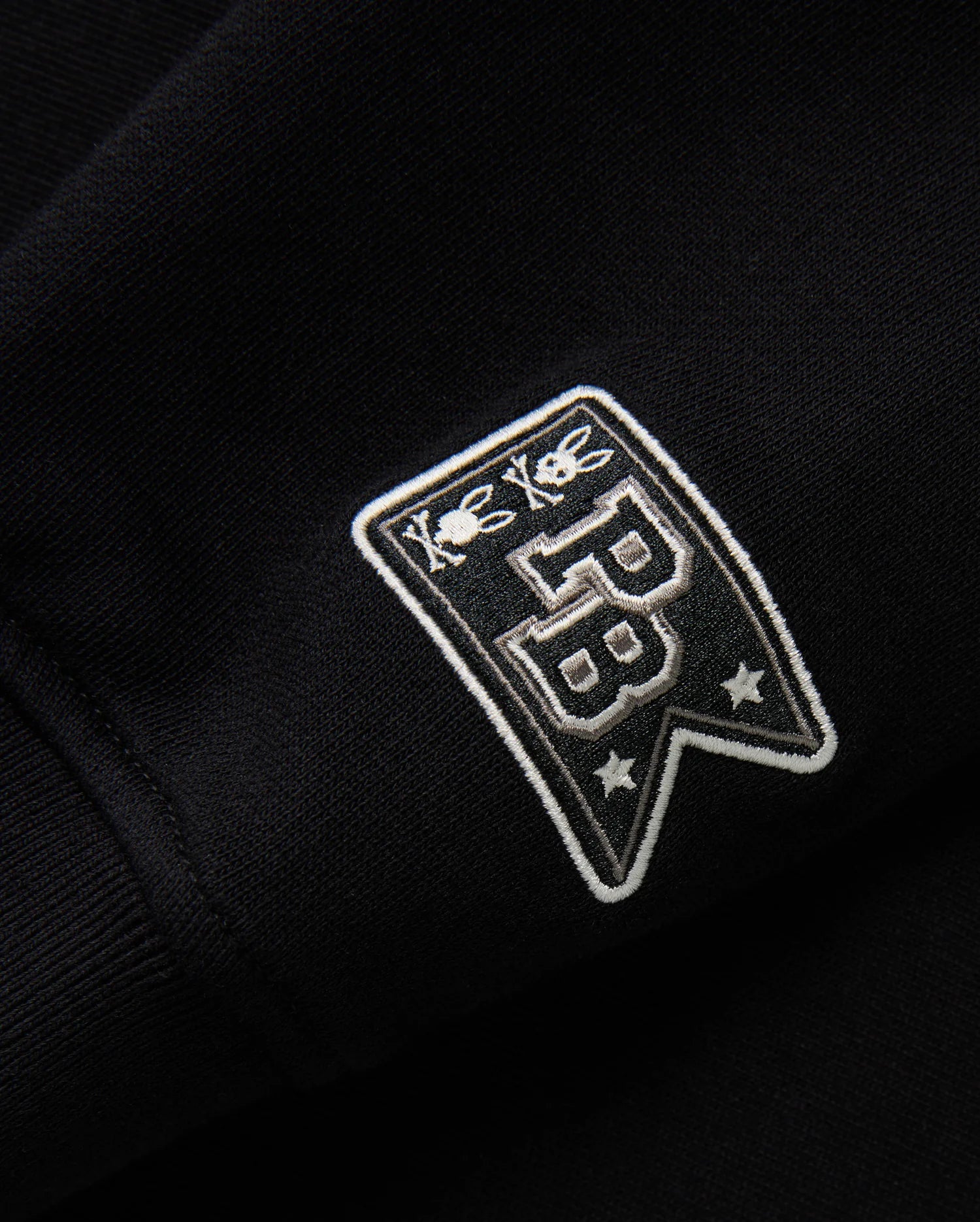 Close-up of the black fabric on the KIDS ZACK VARSITY SWEATSHIRT - B0S880D200 by Psycho Bunny, highlighting a vintage-inspired logo with chenille embroidery. The embroidered patch displays two bees above the letters "PB" and two stars below, all in white thread for a striking contrast against the dark background.