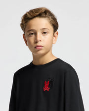 A young person with short, light brown hair is wearing the Psycho Bunny Kids Pierce Long Sleeve Back Graphic Tee (B0T178E200) in black against a neutral background. The tee showcases an embroidered bunny design in red and black on the upper left side. The individual gazes calmly at the camera.