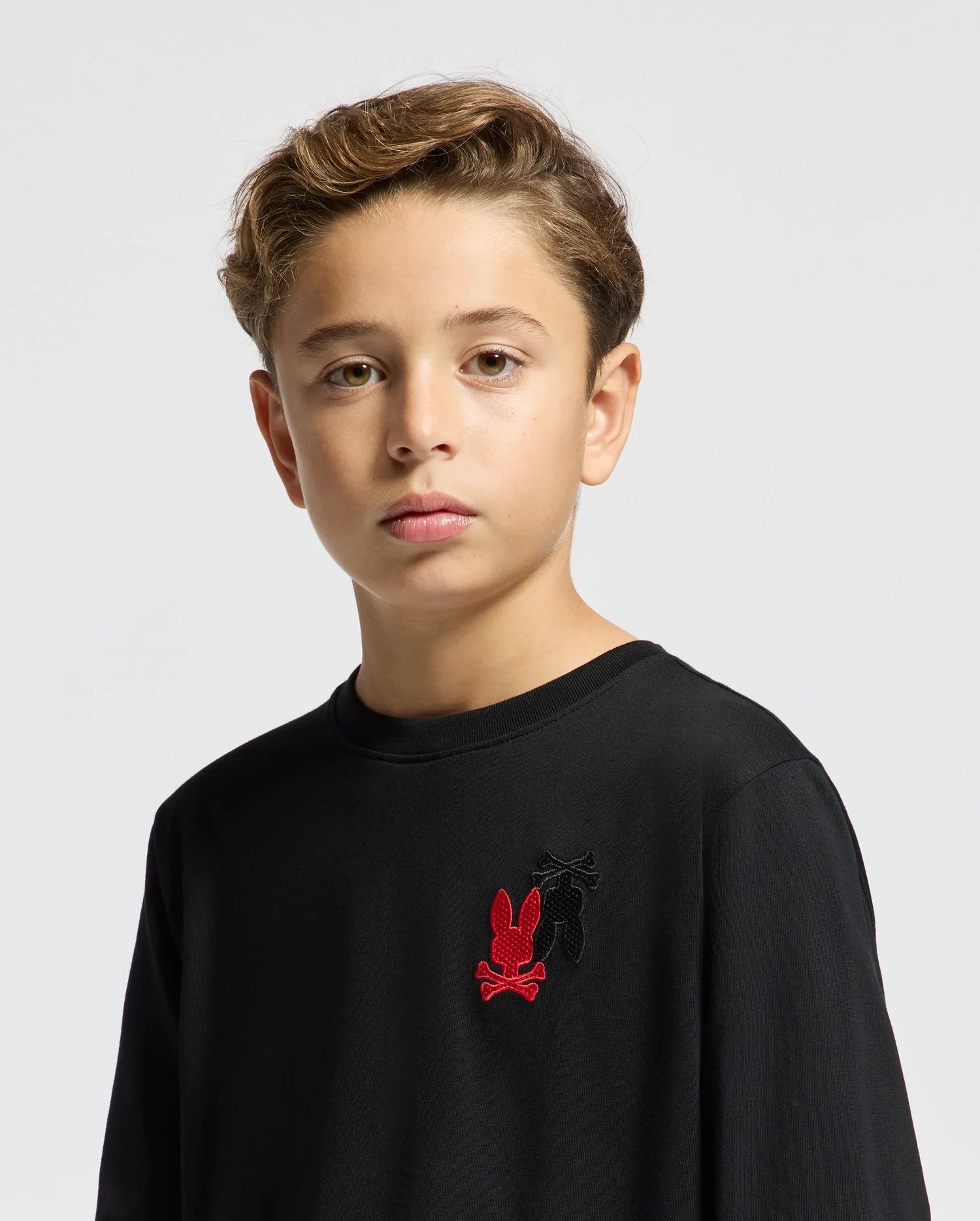 A young person with short, light brown hair is wearing the Psycho Bunny Kids Pierce Long Sleeve Back Graphic Tee (B0T178E200) in black against a neutral background. The tee showcases an embroidered bunny design in red and black on the upper left side. The individual gazes calmly at the camera.