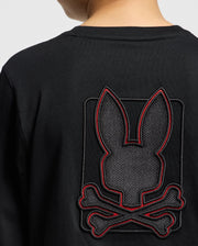 Someone is wearing a black Psycho Bunny Kids' Pierce Long Sleeve Back Graphic Tee, which showcases an embroidered bunny design featuring a stylized bunny head with long ears above crossed bones, all outlined with striking red stitching.