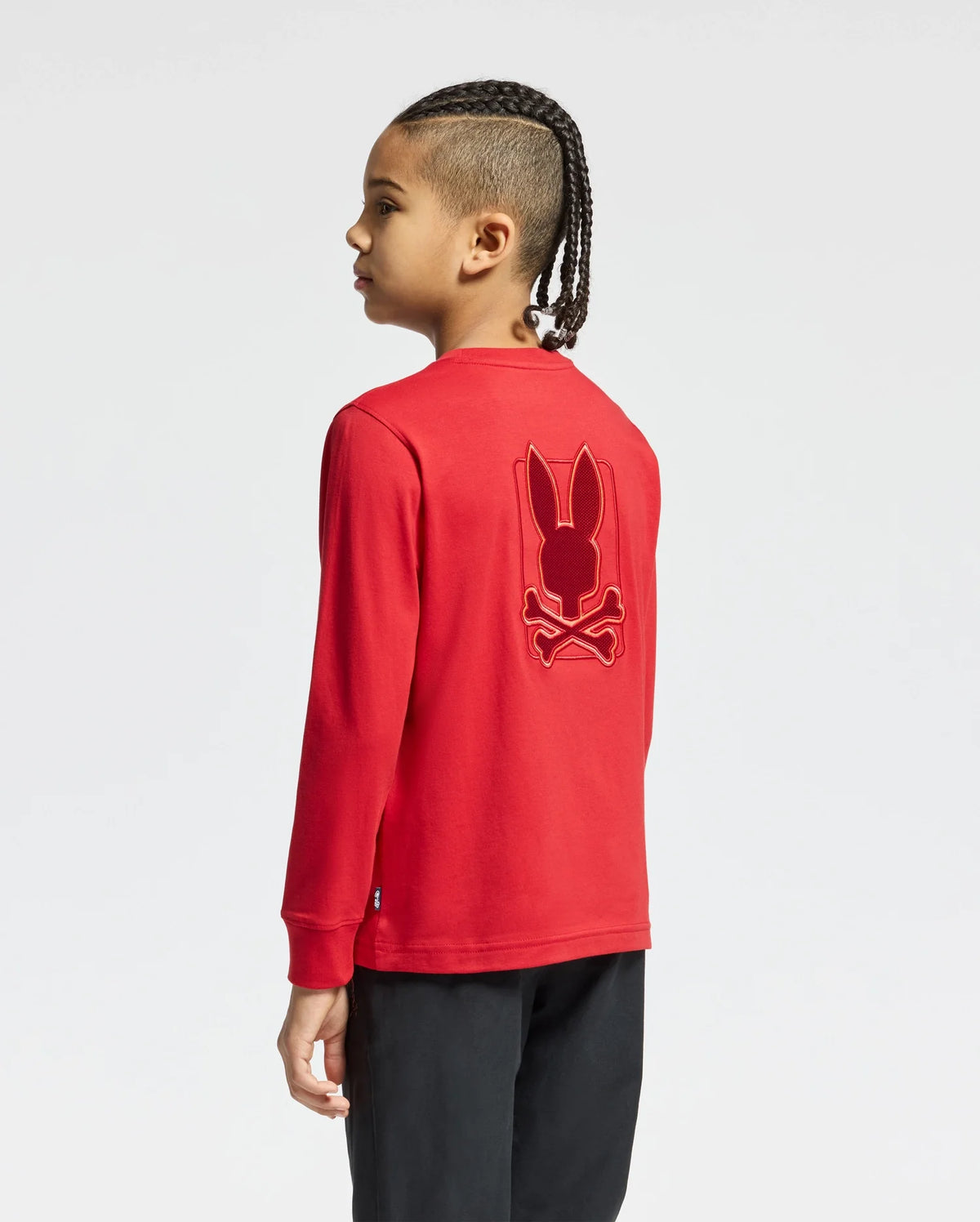 A young individual with braided hair wears the Psycho Bunny KIDS PIERCE LONG SLEEVE BACK GRAPHIC TEE in a vibrant red, featuring an embroidered bunny and crossbones design on the back. Made from luxurious pima cotton, they pose sideways against a plain white background, allowing the design to truly stand out.