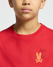 The image features a person wearing the "KIDS PIERCE LONG SLEEVE BACK GRAPHIC TEE - B0T178E200" by Psycho Bunny, which displays embroidered orange and red leaf designs and crossed bone shapes on the chest. The vibrant top, probably crafted from luxurious Pima cotton, is highlighted in the photo to focus on the lower face and upper torso.