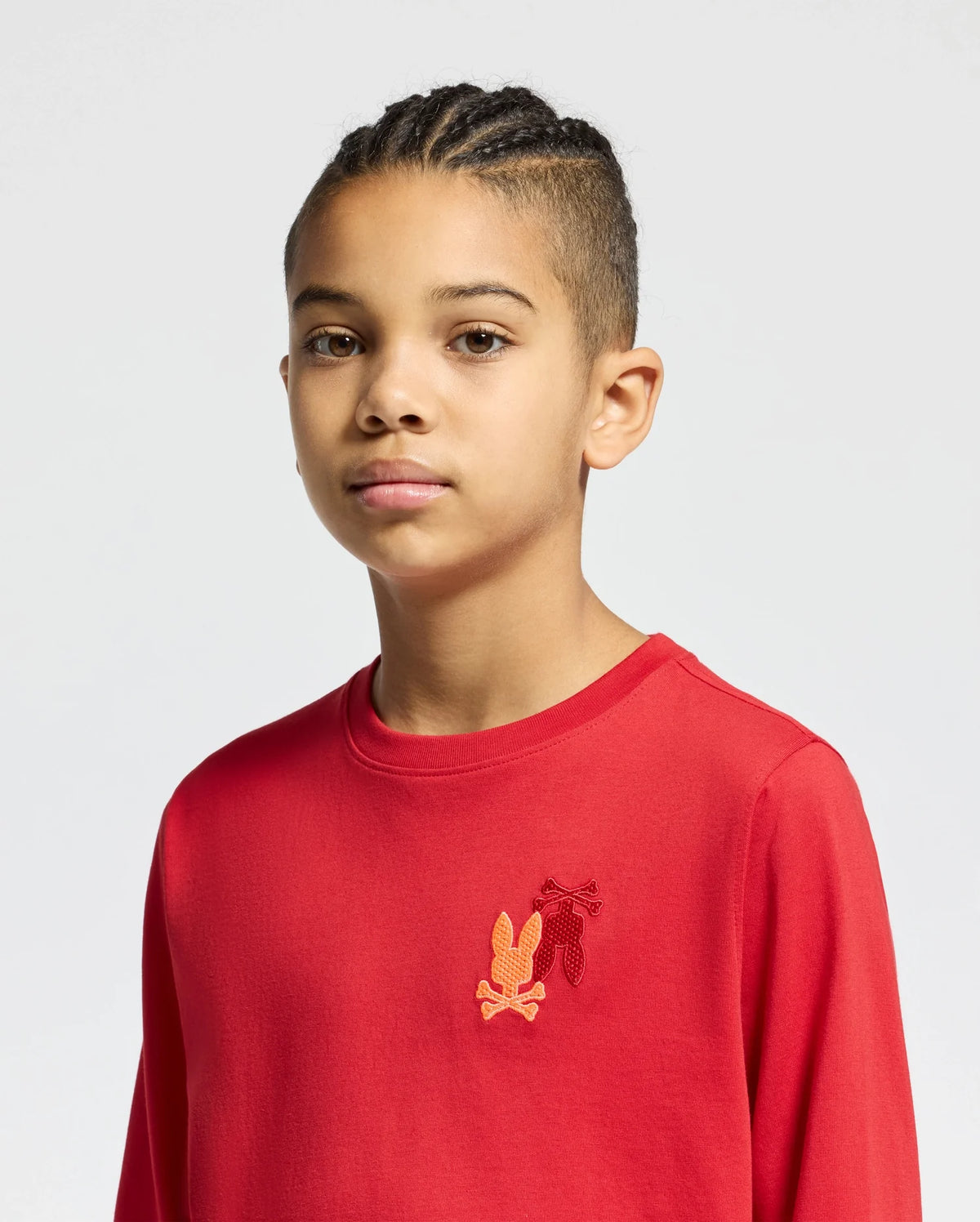 A young person with braided hair is wearing a red Kids Pierce Long Sleeve Back Graphic Tee by Psycho Bunny. The tee, crafted from soft Pima Cotton, features a small embroidered bunny design on the chest against a plain, light-colored background.