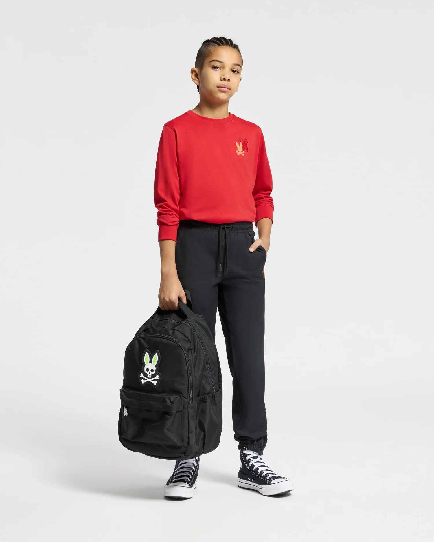 A young individual exudes confidence, donning a red Psycho Bunny Kids Pierce Long Sleeve Back Graphic Tee - B0T178E200 crafted from Pima cotton, paired with black pants. They carry a black backpack featuring an embroidered bunny cartoon design and wear black high-top sneakers, all set against a plain white background.