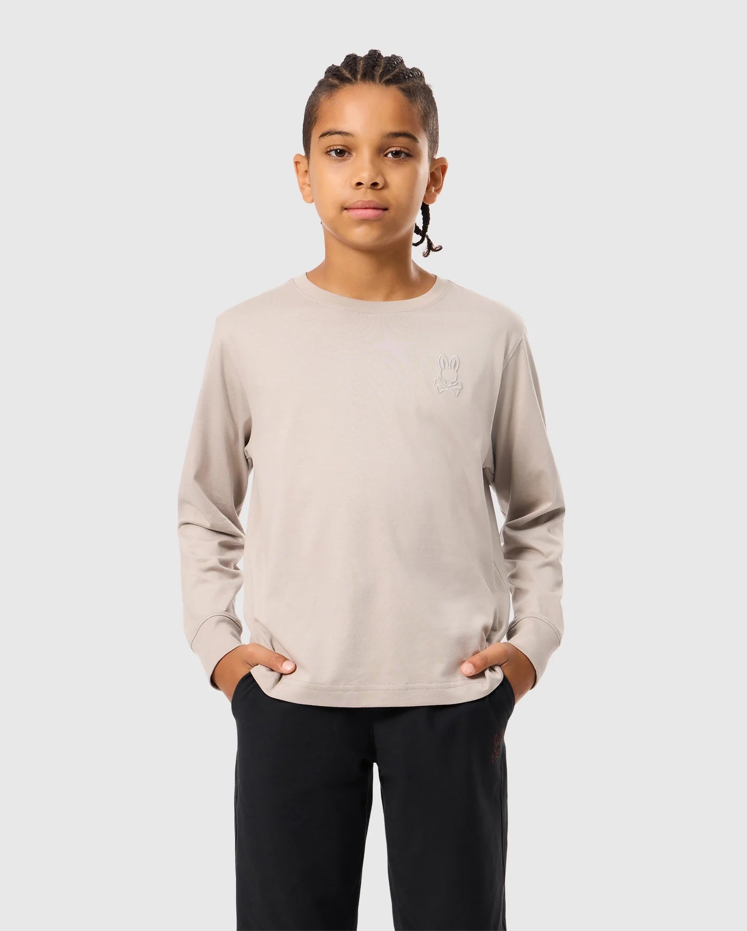 A person with braided hair stands with hands in pockets, wearing a light beige Psycho Bunny Kids Alberton Long Sleeve Tee (B0T867D200) and black pants, against a plain gray background.