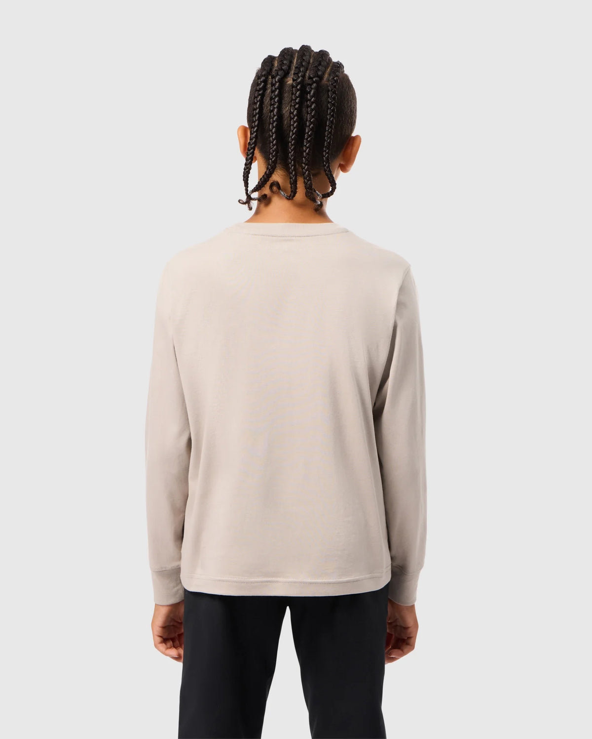 A person with braided hair, wearing the KIDS ALBERTON LONG SLEEVE TEE by Psycho Bunny in a regular fit beige color, paired with black pants, stands facing away against a plain light gray background.