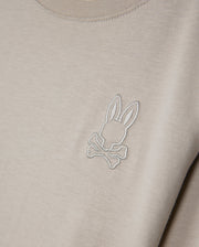 Introducing the Psycho Bunny KIDS ALBERTON LONG SLEEVE TEE - B0T867D200: A close-up showcases this beige, long sleeve tee crafted from soft Pima cotton. It features an embroidered logo of a stylized bunny head above crossed bones, delivering a playful twist on the classic skull and crossbones motif, all while ensuring comfort and style with its regular fit.