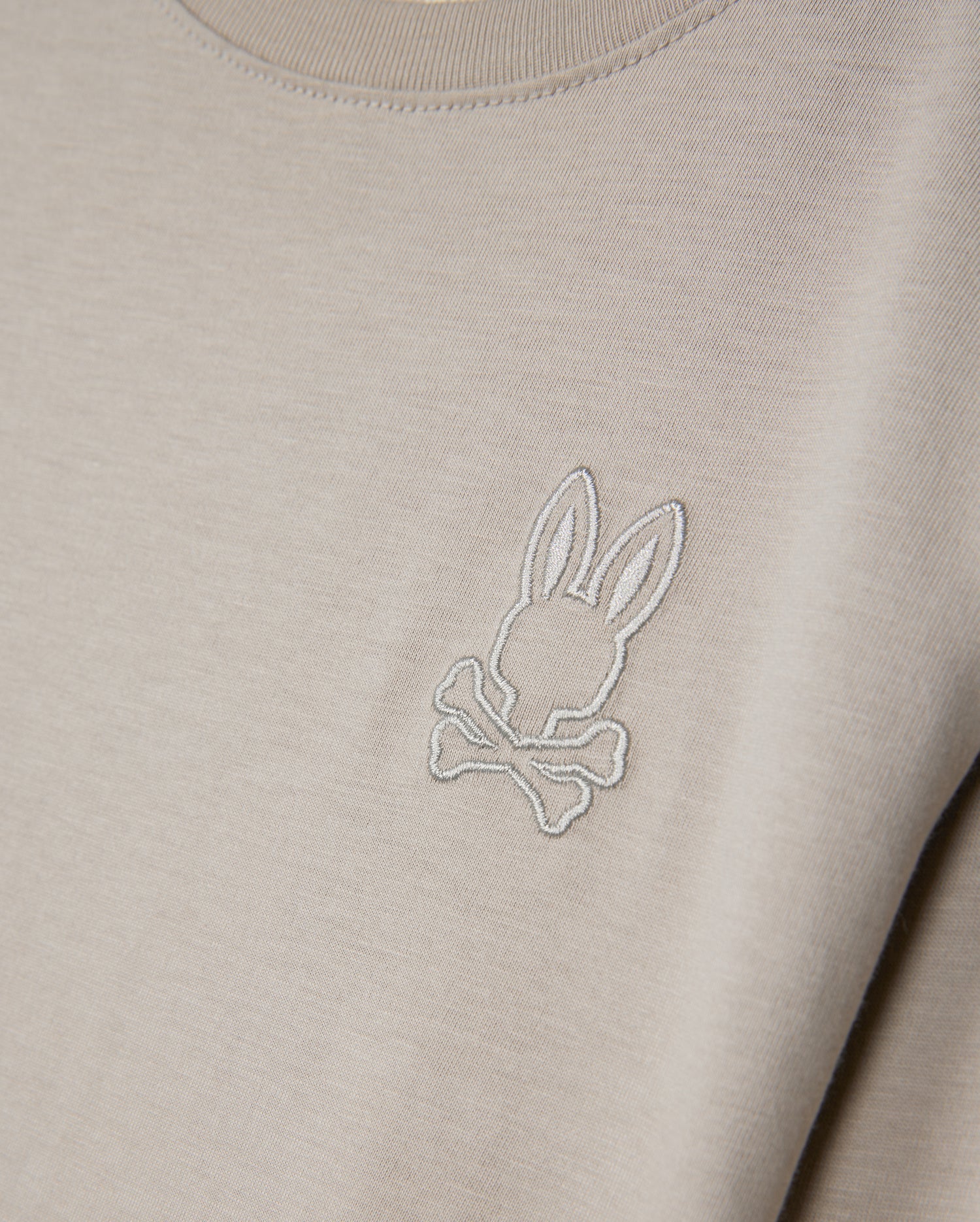 Introducing the Psycho Bunny KIDS ALBERTON LONG SLEEVE TEE - B0T867D200: A close-up showcases this beige, long sleeve tee crafted from soft Pima cotton. It features an embroidered logo of a stylized bunny head above crossed bones, delivering a playful twist on the classic skull and crossbones motif, all while ensuring comfort and style with its regular fit.