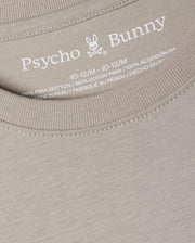 Close-up of a beige KIDS ALBERTON LONG SLEEVE TEE - B0T867D200 by Psycho Bunny, highlighting the brand logo and size details (10-12/M) inside the neckline. This regular fit tee is made from 100% Pima cotton for exceptional softness and quality, crafted in Peru.