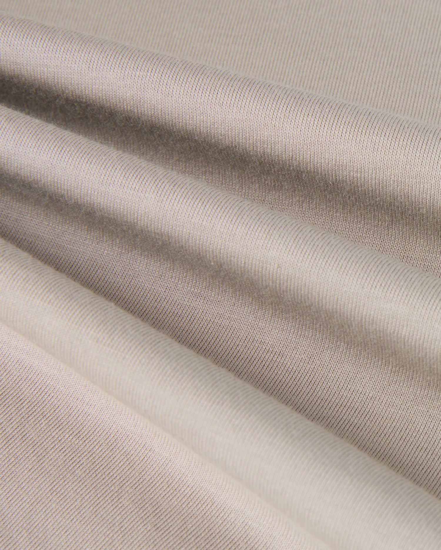 The close-up of the KIDS ALBERTON LONG SLEEVE TEE by Psycho Bunny reveals a beige textured fabric with soft folds and subtle horizontal lines. Made from Pima cotton, this material appears smooth and slightly reflective, creating gentle shadows and highlights across its surface.