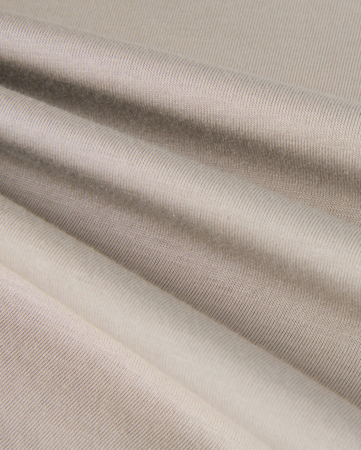 The close-up of the KIDS ALBERTON LONG SLEEVE TEE by Psycho Bunny reveals a beige textured fabric with soft folds and subtle horizontal lines. Made from Pima cotton, this material appears smooth and slightly reflective, creating gentle shadows and highlights across its surface.