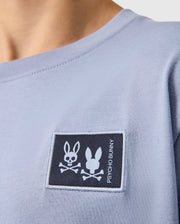 Close-up of someone wearing a Psycho Bunny Kids Monroe Long Sleeve Tee in light blue, featuring a dark blue rectangular chest patch with a bunny skull and crossbones design and "PSYCHO BUNNY," crafted from pima cotton.