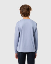 A person with medium-length brown hair stands facing away, wearing a Psycho Bunny KIDS MONROE LONG SLEEVE TEE in light blue and dark pants. The background is plain gray.