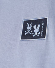 A close-up of the light blue Kids Monroe long sleeve tee in a regular fit features a black patch with Psycho Bunny's logo: two bunny heads with crossed bones—one upright, the other flipped. The brand name is written vertically on the patch for a versatile, cozy style.